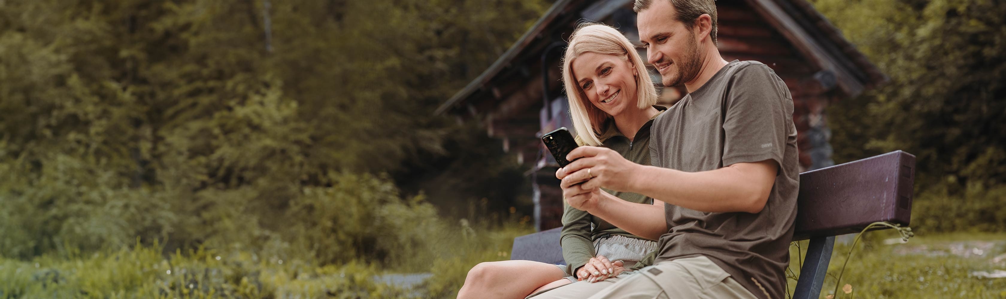 myCONCORDIA – Ein Paar sitzt in Sportbekleidung am Seeufer und schaut auf die myCONCORDIA-App.