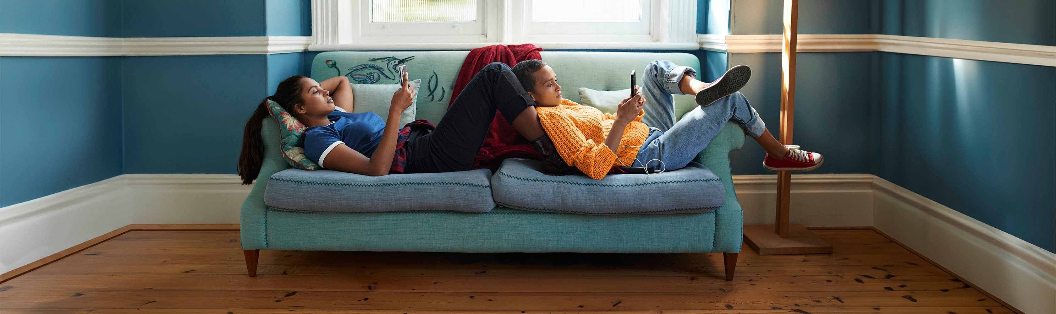 Online Magazin - Zwei Frauen schauen auf ihr Handy, während sie auf einem kleinen, blauen Sofa liegen.