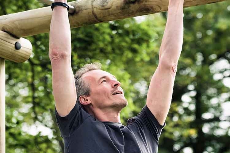 ein Mann mittleren Alters trainiert an einem Hinderniss auf dem Vitaparcours im Wald seine Fitness