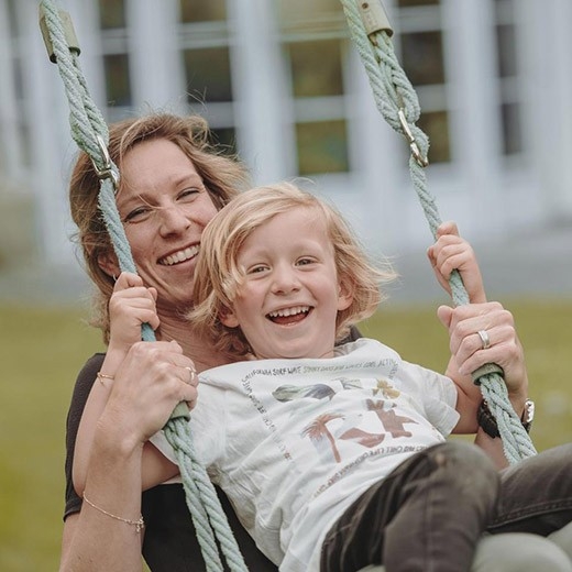 Zusatzversicherungen - Eine Mutter sitzt mit ihrer Tochter auf einer Schaukel, beide lachen fröhlich.