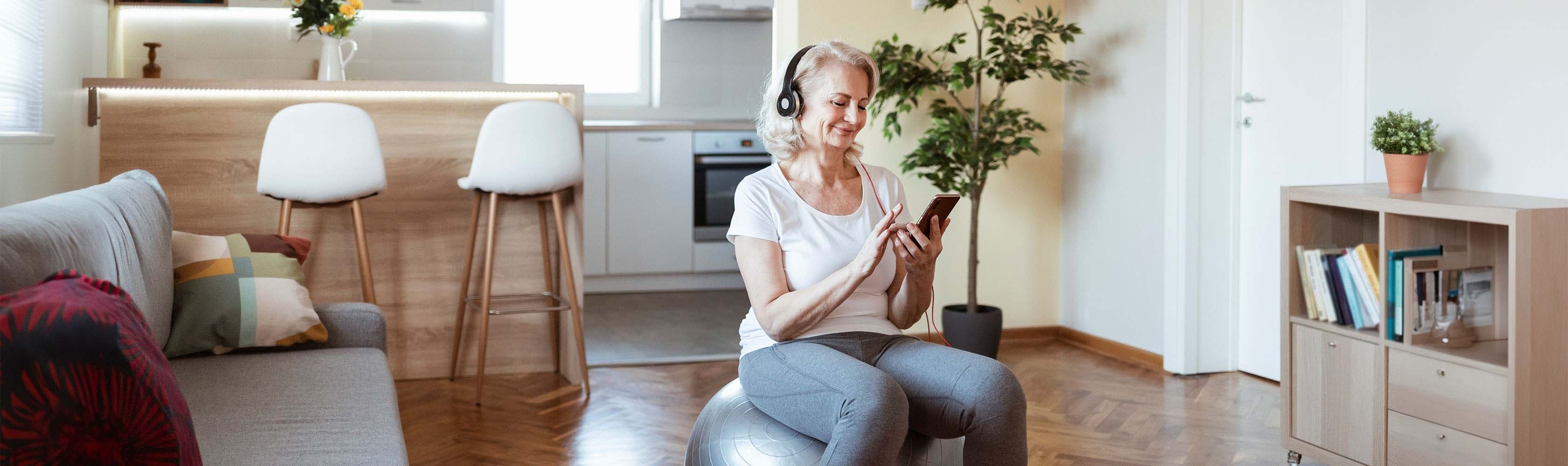 Ältere Frau schaut auf ihren digitalen Begleiter, während sie im Wohnzimmer auf einem Gymnastikball sitzt.