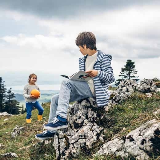 Knabe sitzt auf einem Stein und liest ein Buch. Im Hintergrund steht ein Mädchen mit einem organgen Ball.