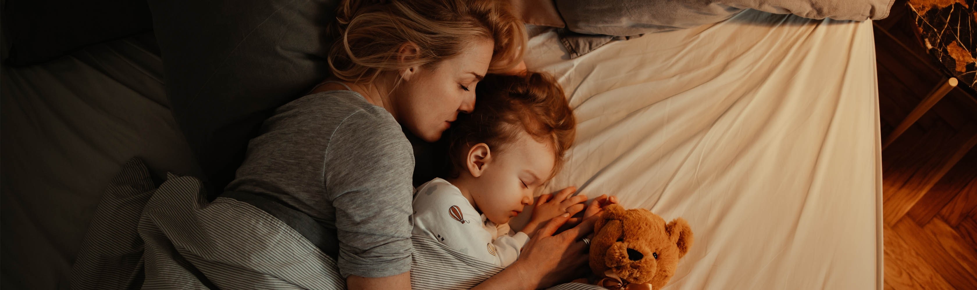Eine Mutter, ein Kind und ein Teddy liegen zusammen im Bett.