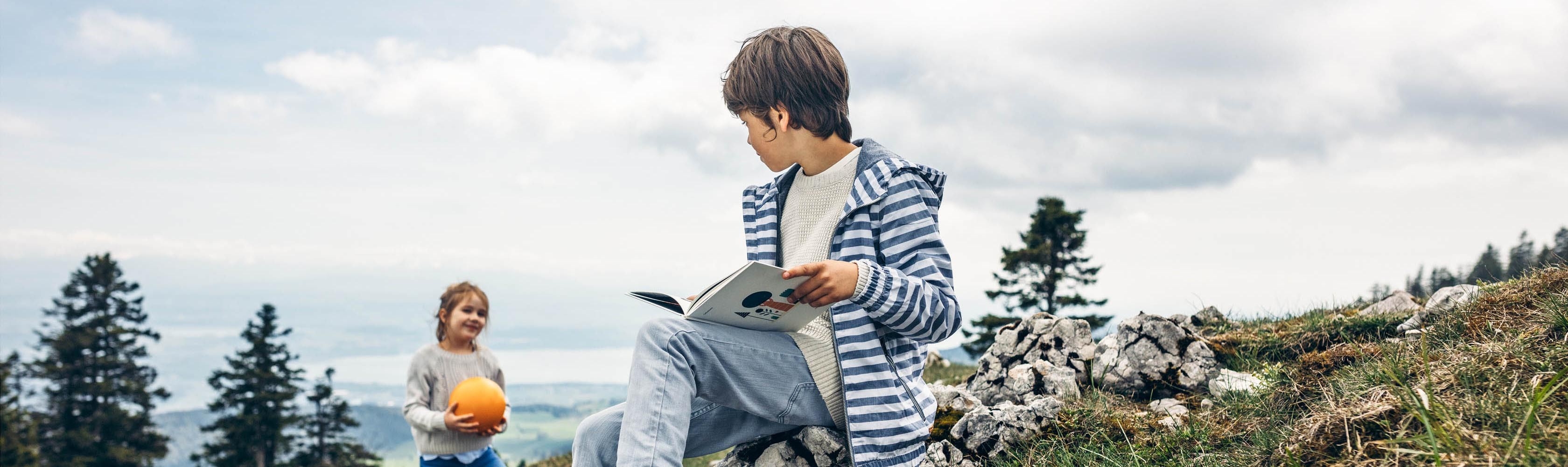 Knabe sitzt auf einem Stein und liest ein Buch. Im Hintergrund steht ein Mädchen mit einem organgen Ball.