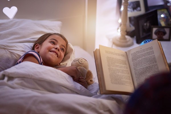 Mädchen liegt im Bett mit einem Teddybär. Ihre Mutter liest aus einem Buch eine Geschichte vor.