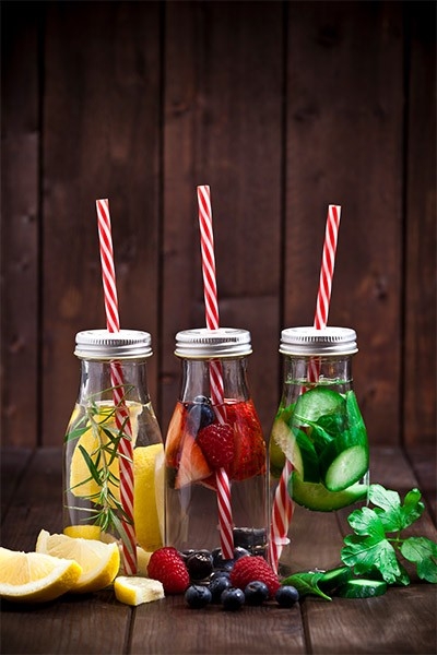 Zu sehen drei Glasflaschen gefüllt mit Wasser und verschiedenen Zutaten wie Zitronenscheiben, Beeren oder Kräutern. Rezepte für herrlich erfrischende, zuckerfreie Getränke.